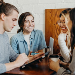 A group of four students sit around a small rectangular table. Several hold travel cups of coffee, and the two in the background smile and laugh. The foremost person looks down at a tablet in their hands.