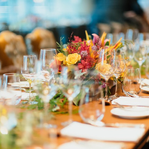 Formal place settings are seen down a long table including three glasses, flatware, a small plate, and napkin at each place. Greenery is strewn down the center of the table with a yellow and red floral arrangement in the center.