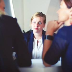 A young woman wearing a blazer sits across from two people, whose backs are to the camera. She appears to listen intently to one of the people across from her.