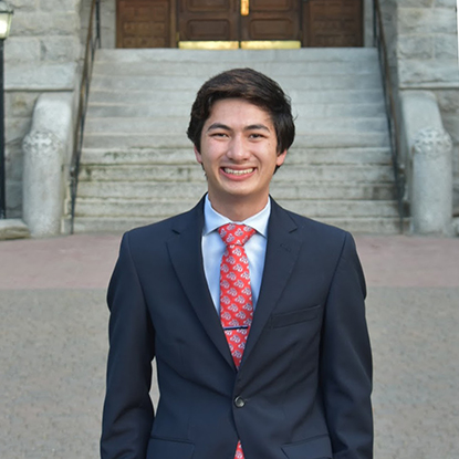 Wearing a navy blazer, blue shirt, and red tie, Charles ("CJ") stands proudly in front of the College Hall steps. 