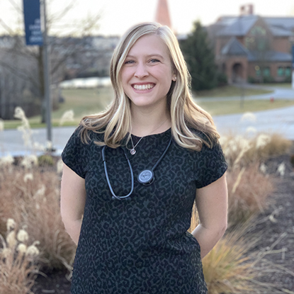 Jordan stands on the Myrtle Woldson Performing Arts Center front steps, with Jundt Art museum just visible to her right. Around her neck, she wears a stethoscope and smiles brightly. 