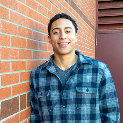 Justis leans against the brick wall of Foley Library, wearing a blue checkered shirt and smiling warmly in the camera. 