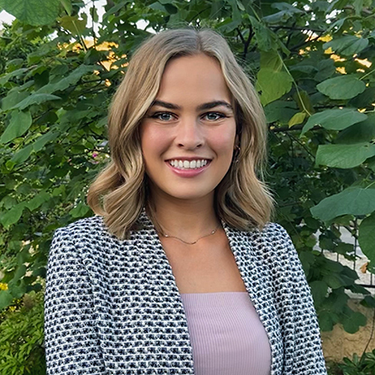 Taylor stands in front of a lush canopy of green branches, wearing a boucle blazer over a pink top. She smiles into the camera. 