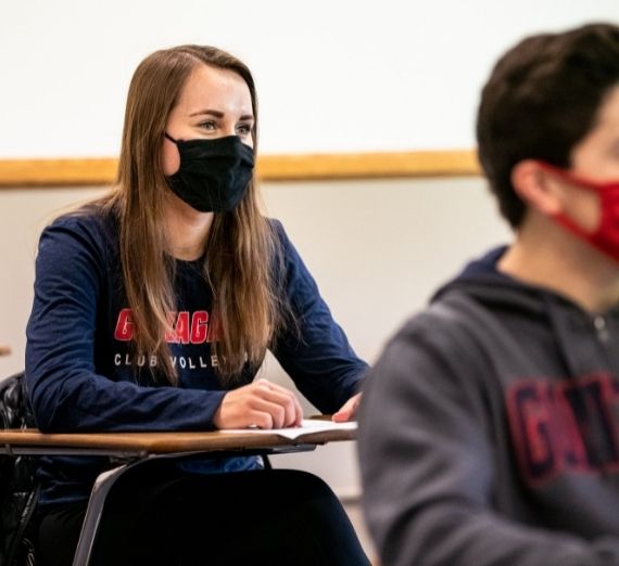 A masked Gonzaga student sitting in class at a desk. Another masked GU student is also in the shot, but blurred to the side.