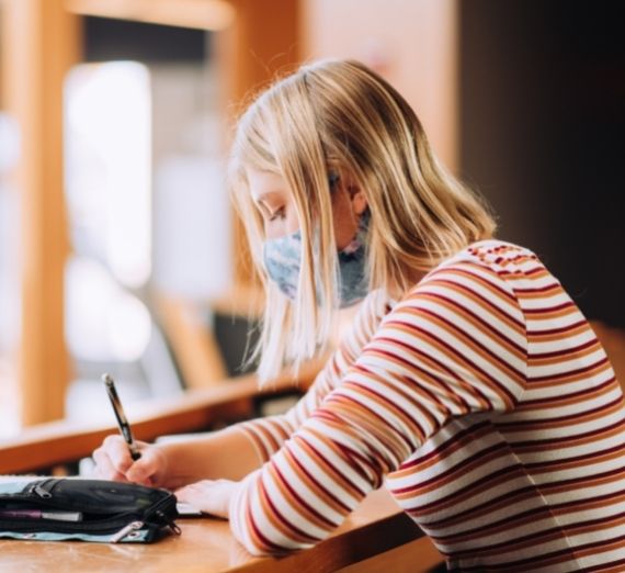 A student sitting indoors writing. She is wearing a blue mask and a red and white striped shirt. Her hair is blonde and the background is blurred.